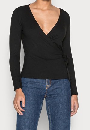 Femme portant un haut cache-cœur noir à manches longues avec un décolleté en V profond, associé à un jean en denim bleu.