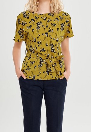 Kvinde iført en sennepsgul kortærmet bluse med sort blomsterprint og sløjfebælte, kombineret med mørkeblå bukser, hænder i lommerne.