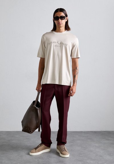 Camiseta beige de manga corta con logo en relieve, pantalones marrón y zapatillas marrón. La figura lleva un bolso tote marrón. Fondo gris.