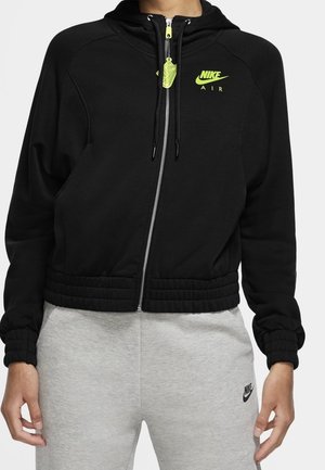 Person wearing black Nike Air zip-up hoodie with neon yellow logo and zipper pull, paired with light gray Nike sweatpants.