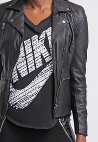 Person i svart läderjacka över svart Nike-t-shirt med stor vit logotyp gjord av liten text, i kombination med svarta byxor med dragkedjedetaljer.