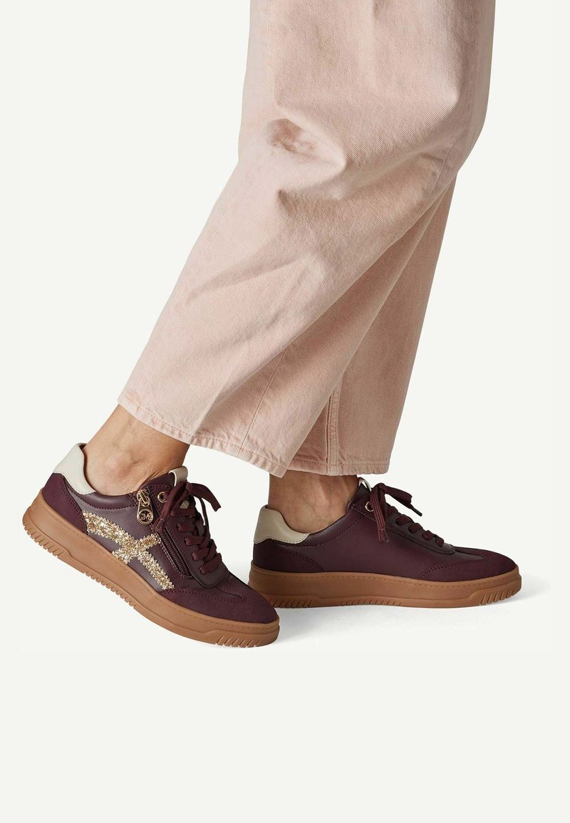 Personne portant un pantalon rose clair et des baskets bordeaux avec des semelles couleur sable et un détail pailleté doré sur le côté, debout sur un fond blanc.