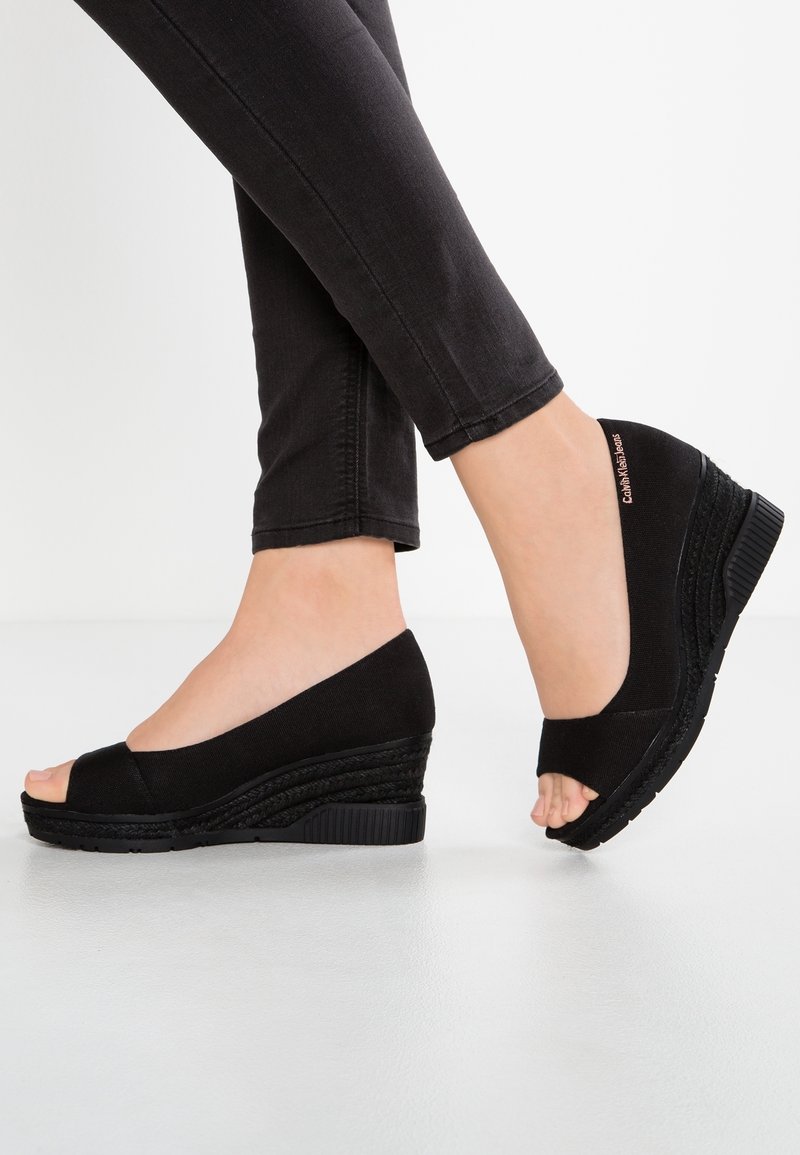 Pieds portant des chaussures compensées noires à bout ouvert, associées à un pantalon noir ajusté sur un fond blanc uni.