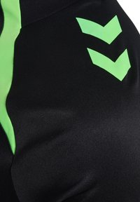 Schwarzer Stoff mit leuchtend grünen Chevron-Logos und grünen Einsätzen, gezeigt ist eine Nahaufnahme eines Details eines Sport- oder Fitnessbekleidungsstücks.