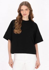 Zwarte, korte mouw top met een ronde halslijn en een oversized pasvorm. Gemaakt van zachte stof, met een subtiele logodetail op de borst.
