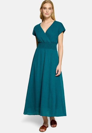 Femme portant une robe turquoise à manches courtes, col en V, longueur cheville avec taille smockée et sandales marron à enfiler, debout devant un fond uni.