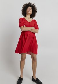 Robe rouge à manches courtes avec un décolleté en V et des boutons décoratifs. Tissu fluide, longueur genou, et présente une coupe décontractée. Portée avec des mocassins noirs.