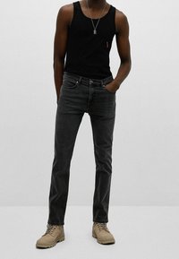 Hombre con camisa sin mangas negra, jeans gris oscuro, botas beige con cordones y un collar de pendiente de plata, de pie con una mano en el bolsillo.