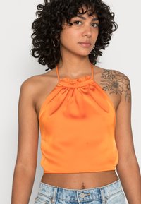 Top cropped halter em satin laranja com um decote ruffled e alças finas, combinado com calções de ganga desgastados de tom azul claro.