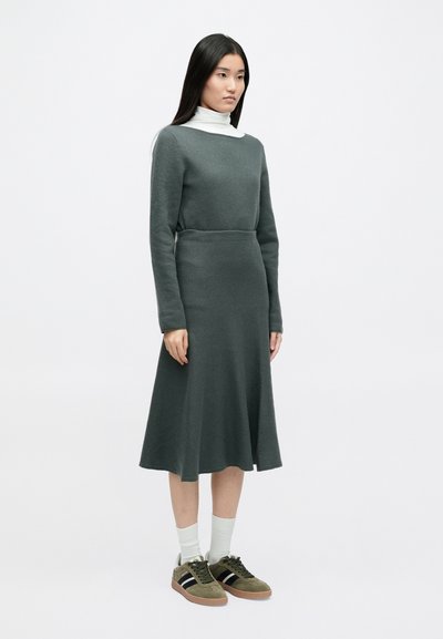 Πλεχτό φόρεμα σε σκούρο πράσινο χρώμα με εφαρμοστό κορσέ και φούστα που φουσκώνει. Συνδυασμένο με σκούρα πράσινα αθλητικά παπούτσια και λευκές κάλτσες μέχρι τον αστράγαλο. Λείο υφή.