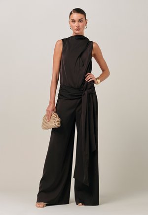 Žena oblečená v čiernom overal s krátkymi rukávmi, širokými nohavicami a bočným pásikom, drží pletené psaníčko, stojí na jednoduchom svetlom pozadí.