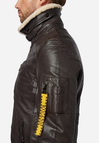 Braune Lederjacke mit einem Schafslederkragen, einem Reißverschlusstaschen an den Ärmeln mit einem gelben Riemen und einer taillierten Passform.