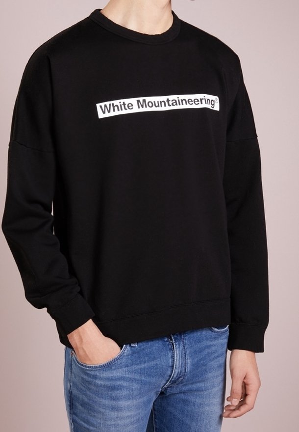 Personne portant un sweat-shirt noir avec le texte « White Mountaineering » sur la poitrine et un jean bleu, debout avec une main dans la poche.