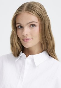 White button-up shirt with a collar, featuring subtle vertical stripes. Smooth texture and a fitted design enhancing the neckline.