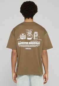 Brązowy bawełniany t-shirt z grafiką na plecach przedstawiającą jedzenie i napis, podkreślający temat „Hangover Breakfast” (Śniadanie po imprezie). Krótkie rękawy.