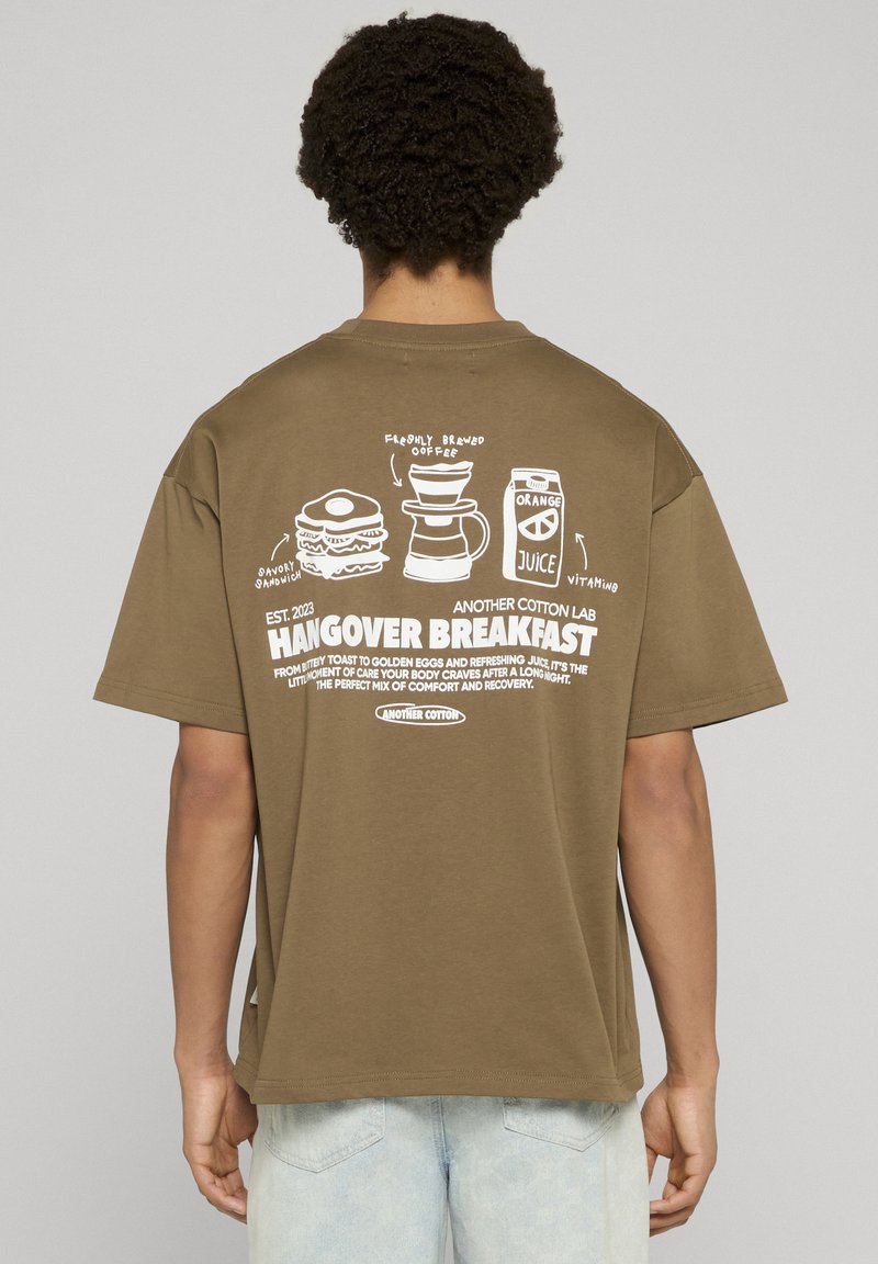 Brązowy bawełniany t-shirt z grafiką na plecach przedstawiającą jedzenie i napis, podkreślający temat „Hangover Breakfast” (Śniadanie po imprezie). Krótkie rękawy.