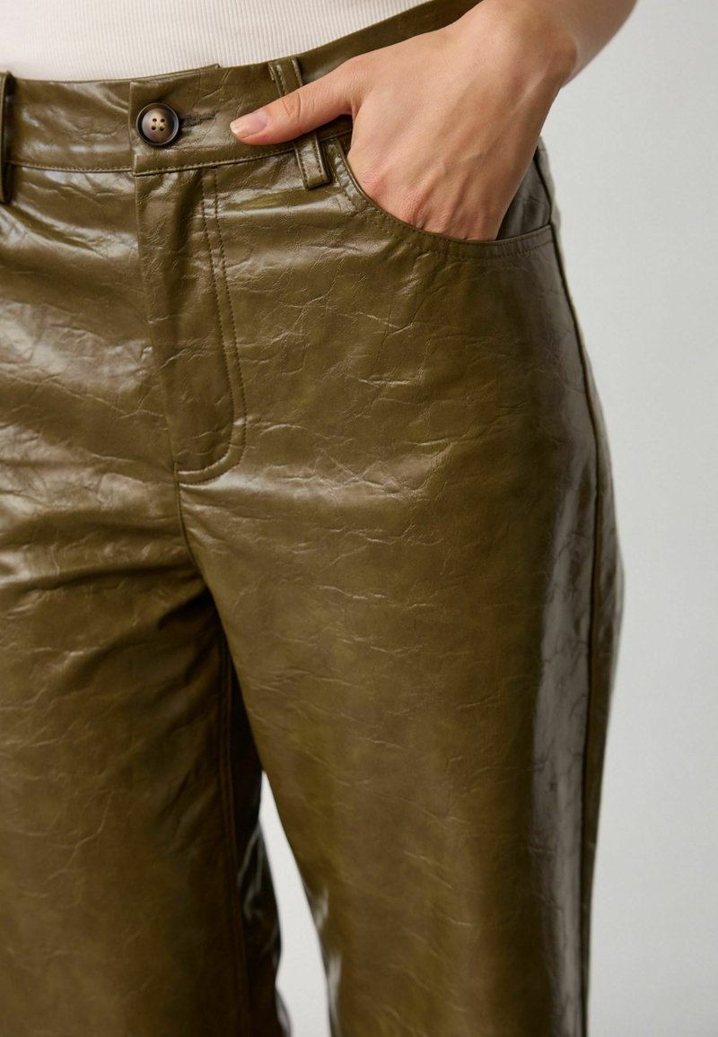 Main reposant dans la poche avant d’un pantalon en faux cuir vert olive avec un bouton et une boucle de ceinture visibles.