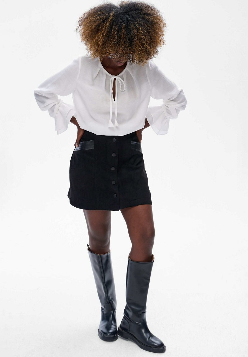Witte blouse met lange mouwen en strikdetail, gecombineerd met een zwarte suède mini rok met frontknopen en zijzakken, afgewerkt met zwarte kniehoge laarzen.
