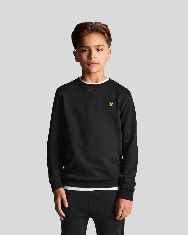 Svart crewneck-tröja i mjukt tyg, med en liten gul logotyp på bröstet och ribbade muddar och nederkant. Stylad med svarta byxor.
