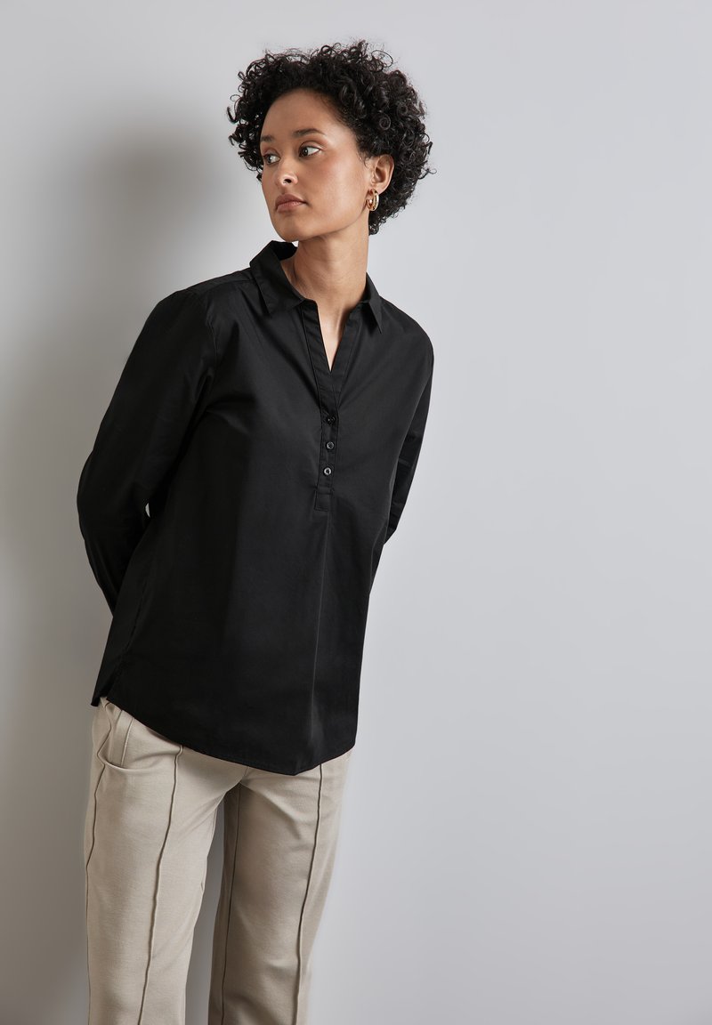 Camicia nera in cotone con colletto, chiusura con bottoni verticali, maniche lunghe e fessure laterali, abbinata a pantaloni beige con piping.