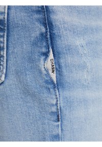 Ljusblå denimjeans med en subtil förstörd textur och en synlig vit etikett med svart text som står "CHASIN" inuti en sidoficka.