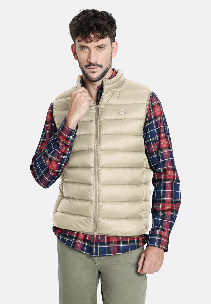 Chaleco acolchado beige con costuras horizontales y cuello alto, usado sobre una camisa de cuadros. El chaleco presenta un pequeño logo y cierre de cremallera.