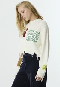Pull court en tricot blanc avec un ourlet effiloché, présentant des imprimés graphiques colorés. Pantalons bleu marine foncé avec une texture lisse et des poches.