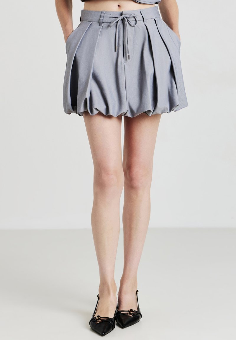Pantaloni corti grigi a plissé con vita regolabile, caratterizzati da una forma a palloncino e tasche laterali. Indossati con tacchi a spillo in vernice nera.