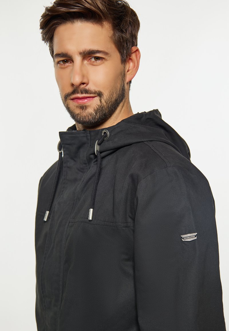 Schwarze Kapuzenjacke aus strukturiertem Stoff, mit silbernen Beschlägen, Kordelzugverschluss und einem Logo-Patch am linken Ärmel.