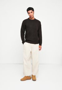 Donkerbruine trui, relaxte pasvorm, geribbelde manchetten; lichtbeige wijde broek; bruine suède schoenen met een textuur. Eenvoudige, moderne uitstraling.