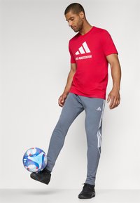 Rød kortærmet t-shirt med hvid tekst "AJAX AMSTERDAM", parret med grå tapered sweatpants med hvide striber og sort fodtøj, svævende en blå fodbold.