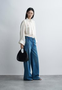 Femme portant une chemise blanche ample et un jean large bleu, tenant un sac à main noir en forme de croissant, debout sur un sol gris contre un mur blanc.