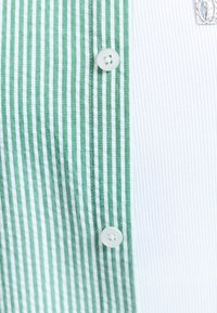 Chemise rayée verticalement vert et blanc avec des boutons blancs, couvrant partiellement un fond en tissu blanc texturé.