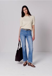 Donna in piedi, indossa una maglietta a maniche corte color crema, jeans blu con cintura leopardata, ballerine leopardate, tiene in mano una grande borsa tote nera.