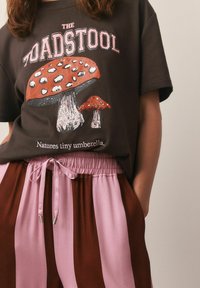 Brun T-shirt med grafik af to røde svampe og teksten "THE TOADSTOOL" ovenfor; parret med elastisk talje striped bukser i pink og brun.