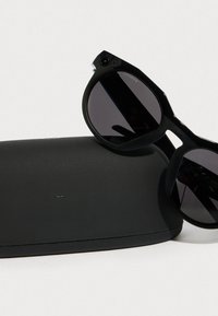 Mustat aurinkolasit, joissa on pyöreät linssit, paksu kehys ja kulmikas muotoilu, asetettuna mustan teksturoidun kotelon viereen.