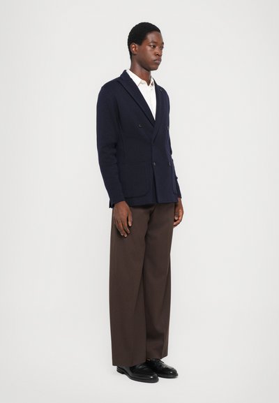 Navy double-breasted blazer in textured fabric with two front pockets, worn over a white shirt, paired with brown trousers and black shoes.