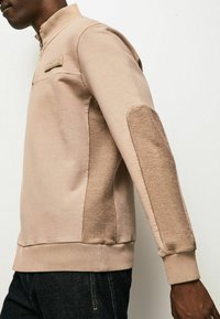 Sudadera con cremallera beige y cuello alto, con parches texturizados en color marrón claro en los codos y puños acanalados. Material suave de mezcla de algodón.