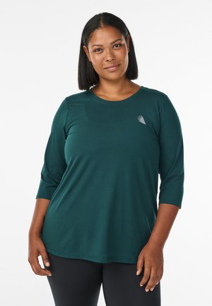 Frau mit glattem schwarzen Haar, die ein dunkelgrünes Langarmshirt und schwarze Leggings trägt, mit neutralem Gesichtsausdruck nach vorne blickend.