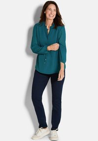 Türkise Bluse mit gerafftem Halsausschnitt, langen Ärmeln und Kordelzugdetails, kombiniert mit dunkelblauen Skinny Jeans und weißen Sneakers.