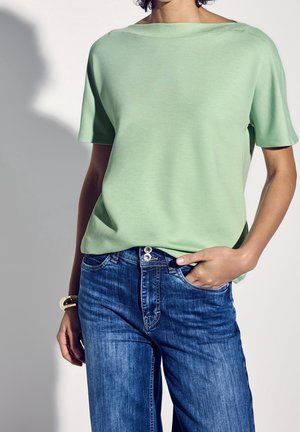 Kobieta ubrana w jasnozielony krótki top z krótkim rękawem i niebieskie dżinsy, z jedną ręką w kieszeni i bransoletką na nadgarstku.