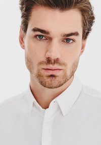 Biała koszula z kołnierzykiem i detalami guzików, zbliżenie na męską twarz o jasnej karnacji, jasnobrązowych włosach i niebieskich oczach na neutralnym tle.