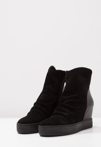 Botines de ante negro con tacón de cuña, que cuentan con cierre de cremallera, textura suave y diseño de material gathered para mayor detalle.