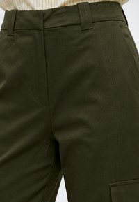 Gros plan d'un pantalon vert foncé avec passants de ceinture, coutures latérales, et vue partielle d'une chemise claire rayée rentrée.