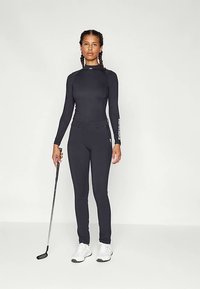 Μαύρο αθλητικό κορμάκι μακρυμάνικο με ψηλό γιακά, σε συνδυασμό με εφαρμοστά μαύρα παντελόνια. Διαθέτει ύφασμα που απομακρύνει την υγρασία και υφή τελειώματος.