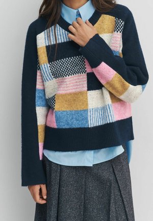 Kvinde iført flerfarvet patchwork-sweater over lyseblå skjorte med krave og mørkegråt skørt, hånd der justerer sweaterens krave.