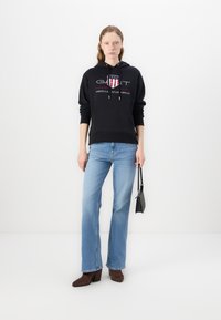 Black hooded sweatshirt with logo and drawstrings, paired with light blue flared jeans and brown heeled boots. Model holds a small black bag.