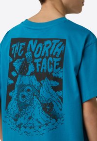 Turkizno turkizna bombažna majica z velikim črnim grafičnim potiskom na hrbtni strani, ki prikazuje gore, oblake in lik z velikimi očmi.