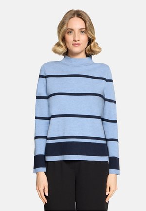 Kvinde med skulderlangt blondt hår iført en lyseblå sweater med mørkeblå vandrette striber og sorte bukser, stående foran en ensfarvet baggrund.