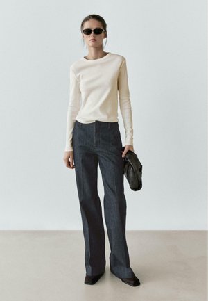 Beiges Langarmoberteil, dunkle Denim-Hose mit weitem Bein und schwarze Sonnenbrille. Hält eine schwarze Tasche mit glatter Oberfläche und maßgeschneidertem Schnitt.